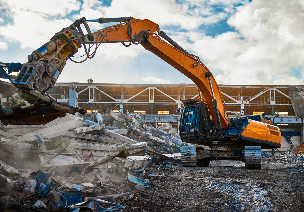 Modern excavator with hydraulic scissors cuts armored concrete beams working at abandoned industrial building demolition site