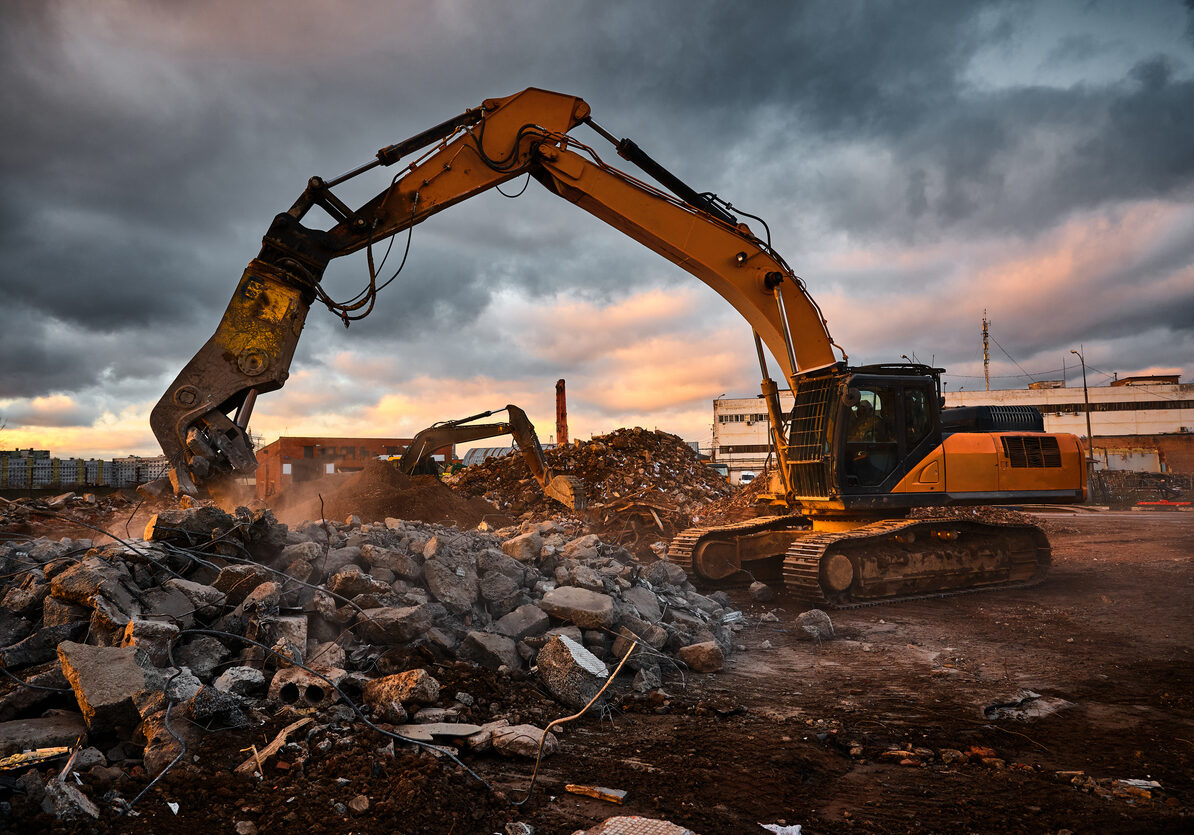 Modern crusher on excavator rigging dismantles reinforced concrete beam for recycling at demolition site at sunset light closeup